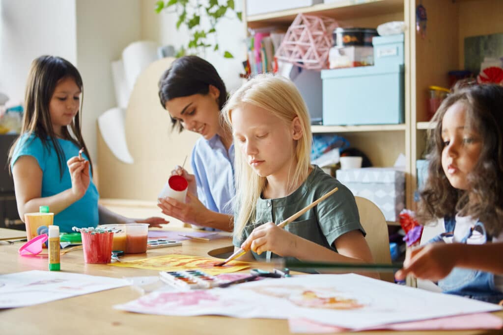 Sweet girls and young teacher painting cute pictures together while sitting at table during art lesson in school